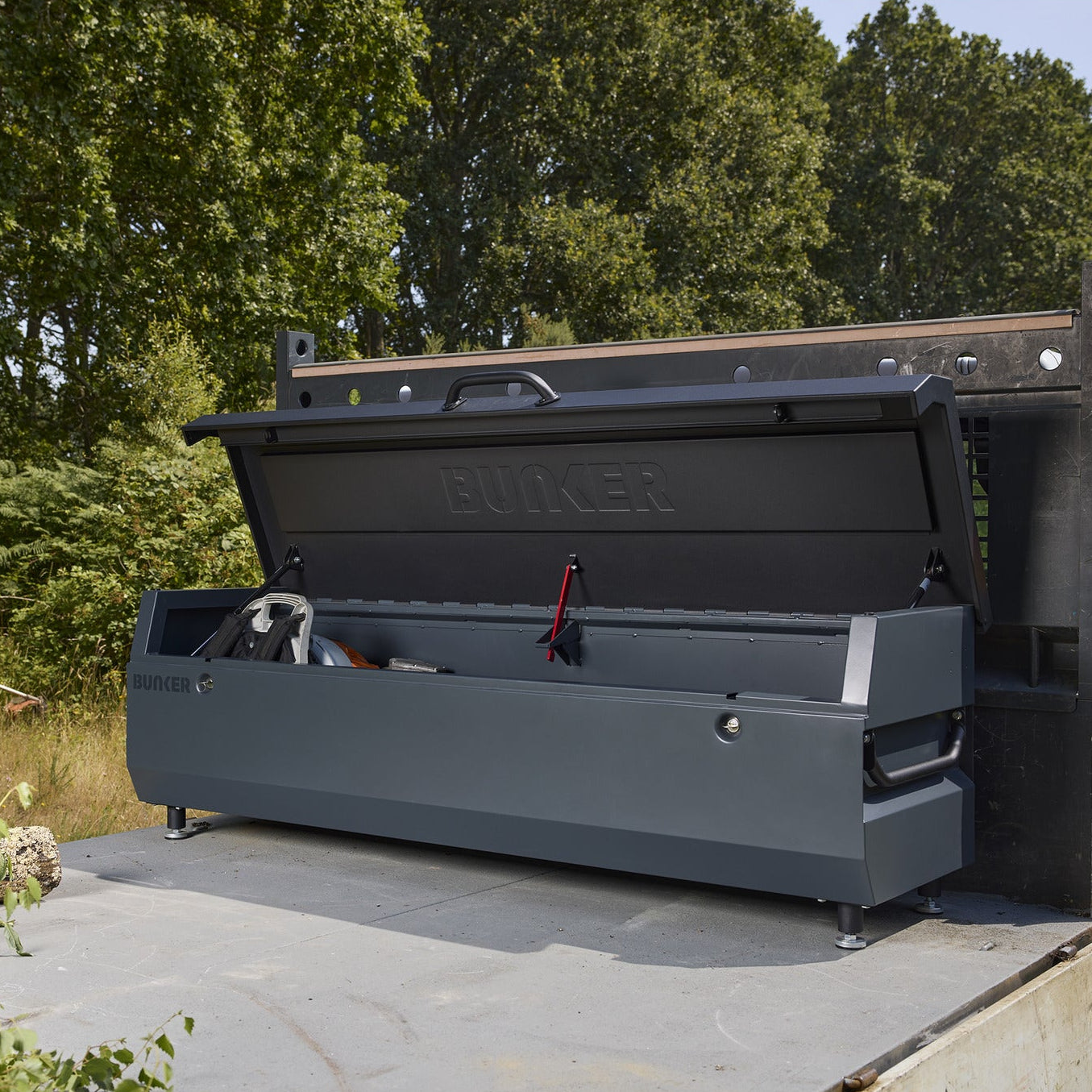 A large dark gray Bicker toolbox mounted on the back of a white utility truck, with a person using a chainsaw in a grassy outdoor area with trees in the background.